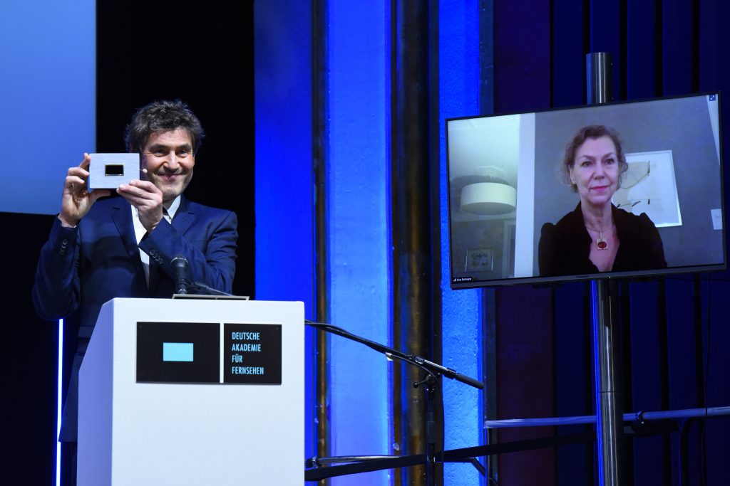 Wir sehen eine Bühne, links Laudator Stephan Szasz und rechts einen Monitor mit der Preisträgerin