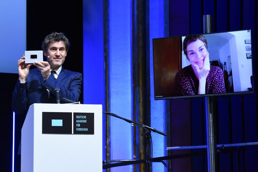 Wir sehen eine Bühne, links Laudator Stephan Szasz und rechts einen Monitor mit der Preisträgerin