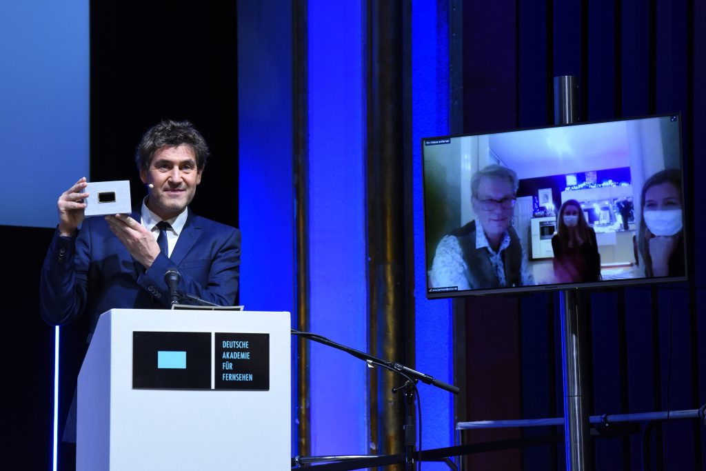 Wir sehen eine Bühne, links Laudator Stephan Szasz und rechts einen Monitor mit den Preisträgern