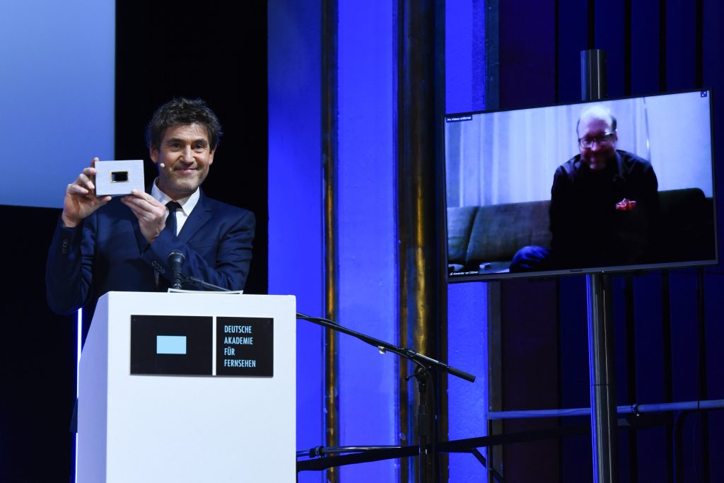 wir sehen eine Bühne. Links den Laudator Stephan Szasz, rechts einen Bildschirm mit dem Preisträger.