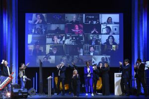 Wir sehen Moderatoren, Band, Laudatoren auf der Bühne vor einer Leinwand. Auf der Leinwand sind die Preisträger 2020 zu sehen